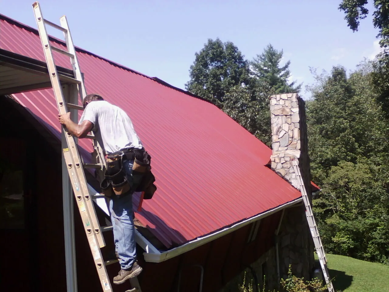 Roofer on a ladder beside a red metal roof and stone chimney — Soffit & Fascia Repair by Roofing Brothers in Usaf Academy